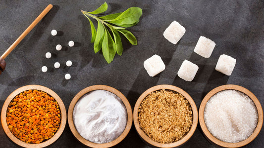 Assorted sweeteners spread on a black tabletop, including stevia leaves, sugar cubes, brown sugar, granulated sugar, erythritol powder, bee pollen, and artificial sweetener tablets