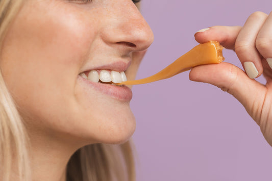 Picture of a young smiling woman biting one side of a caramel, while pulling the other side with her outstretched hand, causing the caramel to stretch