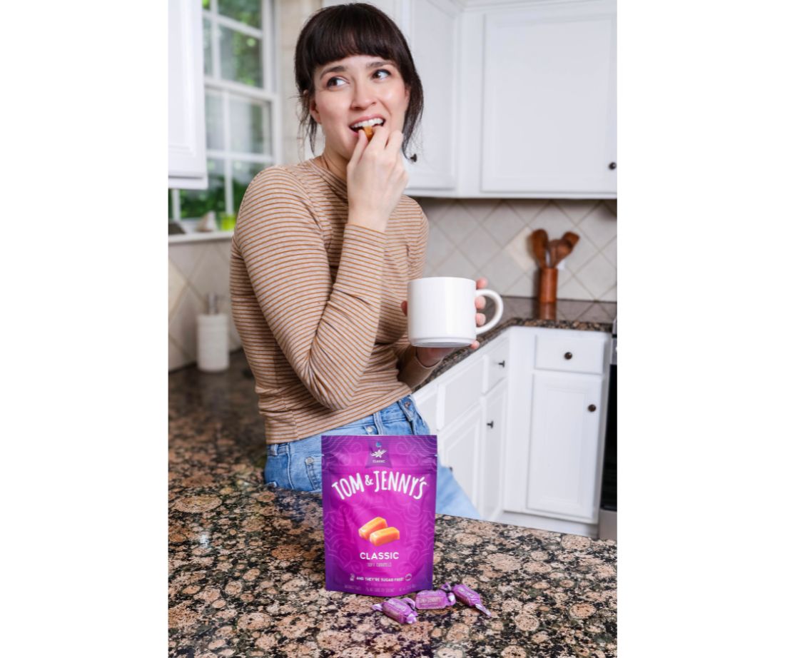 Image showing a woman leaning restfully against a kitchen counter with a large mug in one hand and the other hand is holding onto the caramel she is biting into there is a bag of classic caramels in front of her.