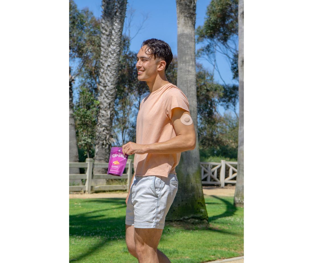 Image showing a young adult man walking in a park holding a bag of classic caramel and he has a continuous glucose monitor visible on his arm.