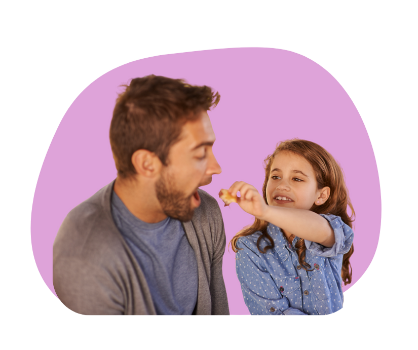 Image of young daughter holding a piece of food towards her dad who has mouth open to eat