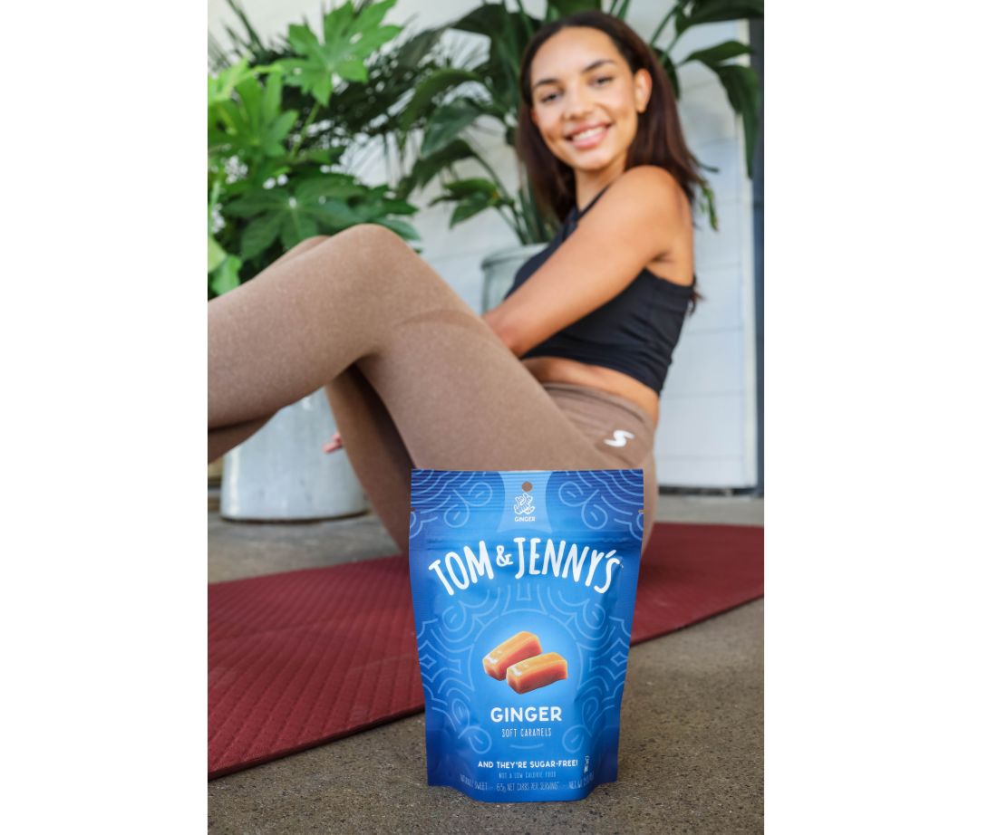 Image showing a young adult woman sitting on a yoga mat smiling in the background with a bag of ginger caramel in the foreground.