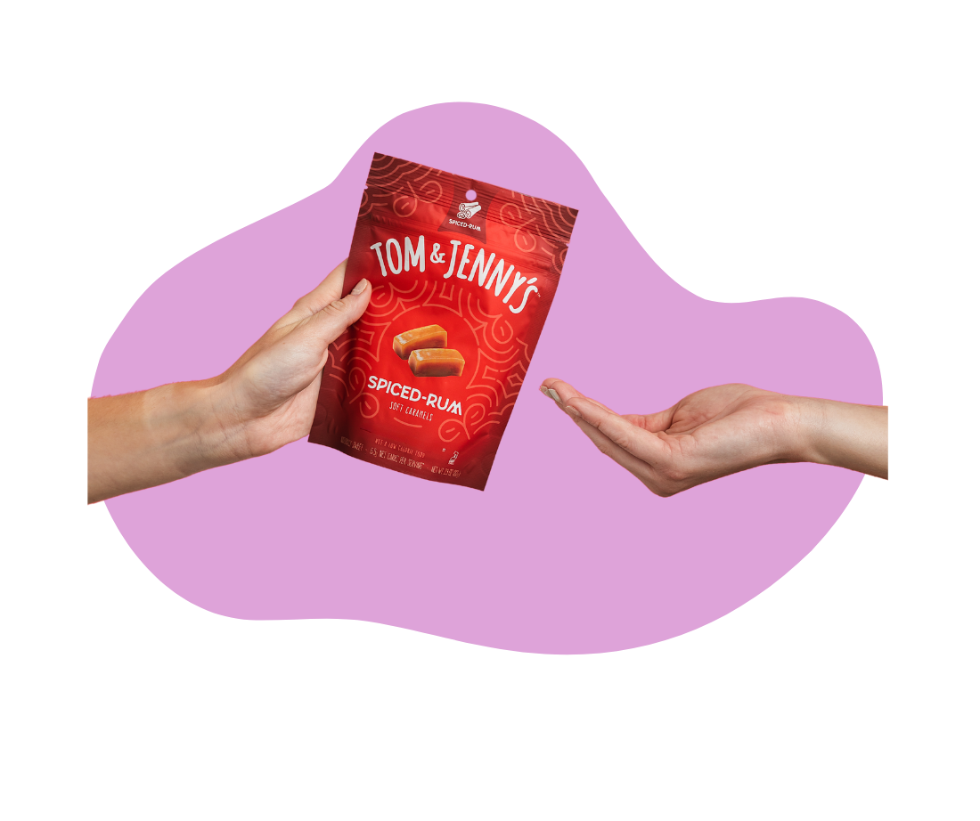 A person's hands are shown holding a red bag of Tom & Jenny's Spiced Rum caramels against a light purple abstract background shape.