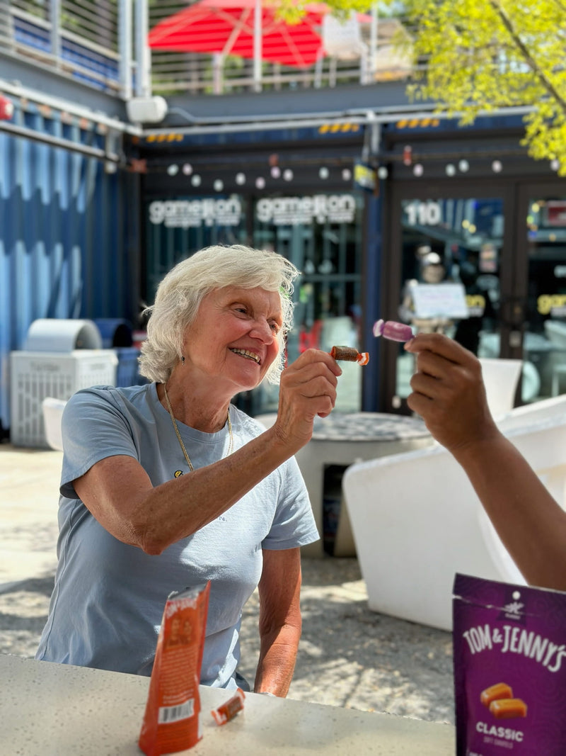 A picture of an older woman smiling and holding up her chocolate Tom & Jenny's, about to "cheers" it with someone else's piece of candy