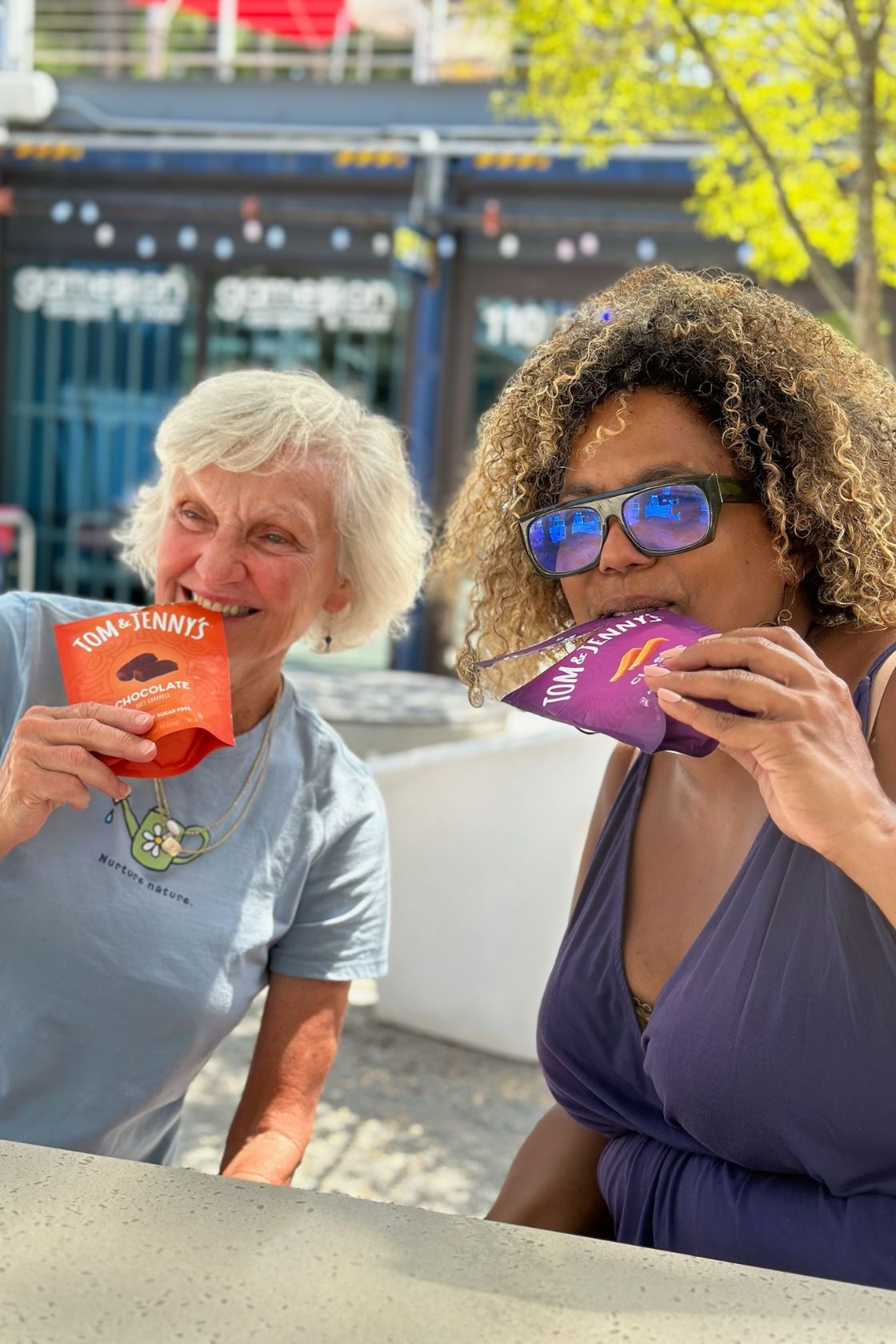 Image of two women playfully biting the corner of bags of Tom & Jenny's