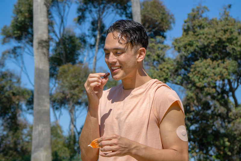 Picture of a young man walking outside with trees behind him. He is snacking on Chocolate Tom & Jenny's and has on a Continuous Glucose Monitor