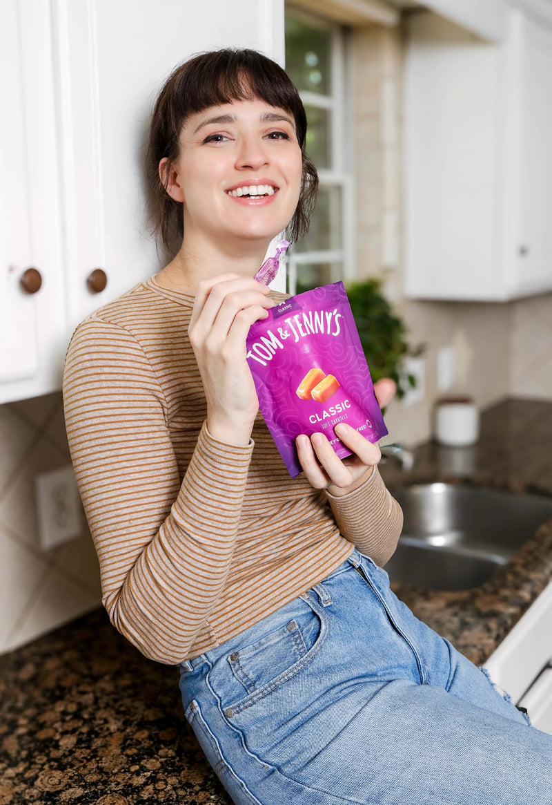 Picture of a young woman sitting on a kitchen counter and grinning while holding a bag of Tom & Jenny's Classic caramels