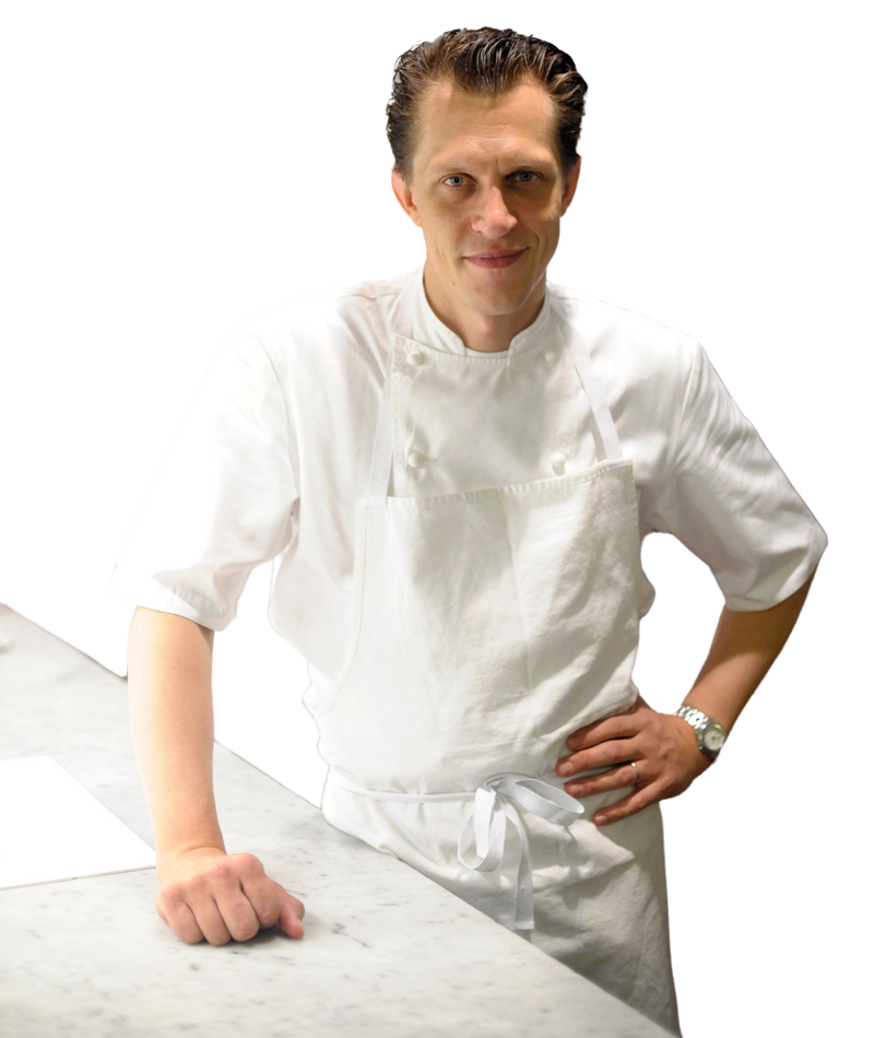 image of chef michael, the james beard winning chef, wearing a white shirt and white chef's apron, standing next to a marble countertop, one hand resting on the counter, the other on his hip, a smile on his face. 